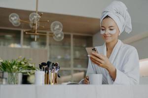 photo d'une jeune femme séduisante porte une serviette de bain sur la tête et une robe de chambre blanche douce tient un téléphone portable aime la communication en ligne applique des patchs hydratants sous les yeux boit une boisson chaude