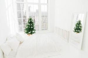 concept de confort, de maison et de réveillon de noël. chambre avec lit moelleux blanc, miroir, grande fenêtre pour la lumière entrant dans la chambre, arbre de nouvel an décoré et échelle. décoration de vacances. photo