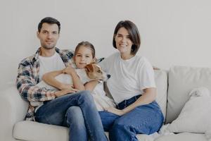 famille amicale pose ensemble au canapé, aime l'atmosphère domestique. le père, la mère, leur petite fille et leur chien de race passent le week-end à la maison, posent dans le salon, ont des expressions de visage heureux photo