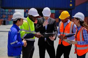 un groupe de travailleurs multiraciaux en uniformes de sécurité et casques travaillent au quai du terminal logistique avec des piles de conteneurs, le contrôle du chargement et la gestion de l'expédition des marchandises, l'industrie du transport de fret. photo