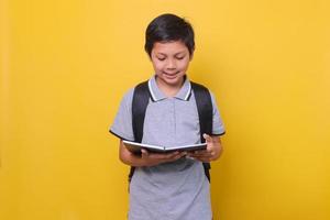 écolier asiatique dans un style décontracté avec sac à dos noir sourit en lisant un livre isolé sur fond jaune. concept de retour à l'école. photo