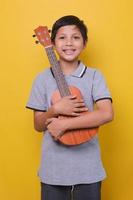 un garçon asiatique heureux sourit et tient un ukulélé. enfant heureux appréciant la musique. éducation, école de musique, cours. style de vie et profiter de la musique. photo