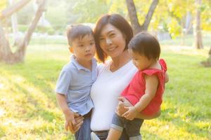 belle jeune mère asiatique portant un petit garçon et une fille dans le parc, femme asiatique heureuse d'avoir un fils et une fille et un enfant câlin, maman aime et embrasse l'enfant ensemble, fête des mères et concept de famille. photo