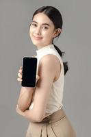 femme asiatique confiante tenant un téléphone montrant un écran vide et regardant la caméra isolée sur fond gris. portrait d'une belle fille en studio. photo