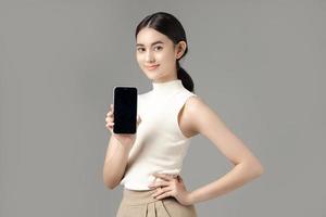 femme asiatique confiante tenant un téléphone montrant un écran vide et regardant la caméra isolée sur fond gris. portrait d'une belle fille en studio. photo
