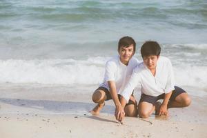 jeune couple asiatique gay souriant dessin romantique en forme de coeur ensemble sur le sable en vacances, homosexuel heureux et amusant avec amour assis sur le sable à la plage en voyage d'été, concept juridique lgbt. photo