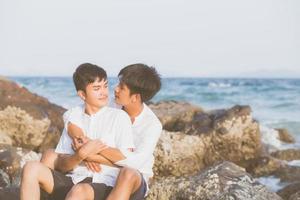 portrait homosexuel jeune couple asiatique assis câlin ensemble sur un rocher ou une pierre sur la plage en été, asie gay faisant du tourisme pour les loisirs et se détendre avec plaisir en vacances en mer, concept juridique lgbt. photo