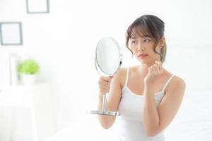 belle jeune femme asiatique regardant le miroir avec un problème d'acné dans la chambre, un problème de beauté sur le visage, un traitement de zit, une fille asiatique est un bouton inquiet et mécontent, des soins de la peau et un concept sain. photo