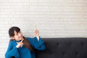 beau portrait jeune femme asiatique pointant avec espace de copie sur fond de béton de ciment, fille souriante assise sur un canapé et montrant l'avenir ou la planification avec un message de vous. photo