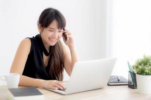 belle jeune femme asiatique indépendante souriante travaillant sur un ordinateur portable et parlant d'un téléphone intelligent mobile au bureau avec un professionnel, une fille utilisant un ordinateur portable, un concept d'entreprise et de style de vie. photo