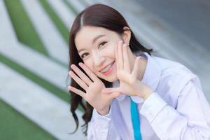 belle étudiante asiatique de lycée dans l'uniforme scolaire se tient debout et sourit joyeusement avec des accolades sur ses dents pendant qu'elle met sa main sur le visage en toute confiance avec le bâtiment en arrière-plan. photo