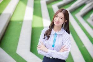 belle étudiante asiatique de lycée dans l'uniforme scolaire se tient debout et sourit joyeusement avec des accolades sur ses dents pendant qu'elle traverse le bras en toute confiance avec le bâtiment en arrière-plan. photo