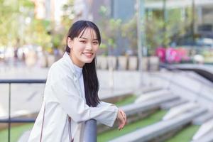 une jeune femme d'affaires asiatique confiante qui porte une chemise blanche et un sac à bandoulière sourit joyeusement alors qu'elle se tient sur le chemin du bureau pour travailler dans la ville. photo