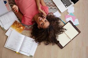 vue de dessus intérieure d'une belle jeune femme bouclée à la peau foncée, faisant une pause avec les devoirs et donnant un appel à un ami, allongée sur un tapis avec des livres et des notes photo