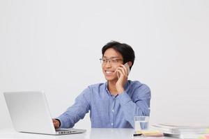 heureux jeune homme d'affaires asiatique réussi dans des verres et une chemise bleue parlant sur un téléphone portable travaillant avec un ordinateur portable à la table sur fond blanc photo