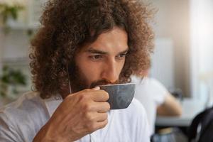 gros plan intérieur d'un homme barbu attrayant aux cheveux bruns bouclés buvant du thé au café, pensant à quelque chose de positif, portant des vêtements décontractés photo