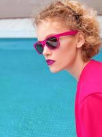 portrait d'une merveilleuse jeune femme bouclée rousse élégante assise en veste fuchsia, lunettes de soleil élégantes et sous-vêtements noirs au bord de la piscine. photo