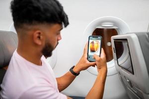 jeune homme latino-américain faisant un appel vidéo sur son téléphone portable, assis dans l'avion près de la fenêtre. photo