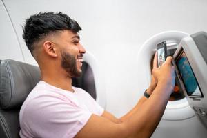 jeune homme latino-américain souriant faisant un appel vidéo sur son téléphone portable, assis dans l'avion près de la fenêtre. photo