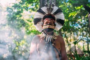 chaman de la tribu pataxo. homme indien âgé portant une coiffe de plumes et un masque facial en raison de la pandémie de covid-19. indien brésilien regardant la caméra photo