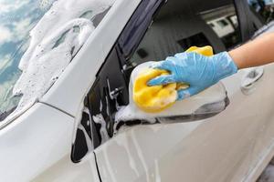 main de femme portant des gants bleus avec une éponge jaune miroir latéral de lavage voiture moderne ou automobile de nettoyage. concept de lavage de voiture photo