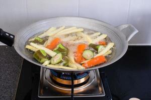 les aubergines, les carottes, les petits épis de maïs ont été transformés en morceaux. et faites-le bouillir dans une casserole de nombreux types de légumes mélangés sont cuits dans une casserole en fer. la casserole en fer fait bouillir des légumes sur la cuisinière à gaz de la cuisine. photo