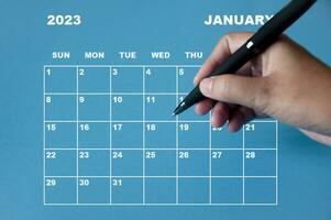 calendrier de janvier 2023 avec main tenant un stylo noir sur fond bleu. photo