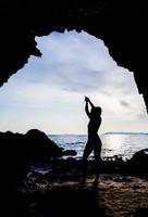 silhouette d'une femme levant les mains pour le yoga ou posant pour ouvrir l'esprit à de belles choses. photo d'une femme pratiquant le yoga debout dans une grotte surplombant la mer au lever ou au coucher du soleil.