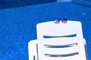 des lunettes de soleil reposent sur une chaise longue blanche sur fond de piscine à l'eau bleue. copie espace photo