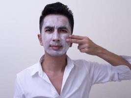 beau jeune homme asiatique appliquant de la crème sur son visage avec un visage souriant, concept de soins de la peau photo