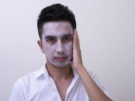 beau jeune homme asiatique appliquant de la crème sur son visage avec un visage souriant, concept de soins de la peau photo