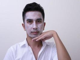 beau jeune homme asiatique appliquant de la crème sur son visage avec un visage souriant, concept de soins de la peau photo