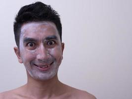 beau jeune homme asiatique appliquant de la crème sur son visage avec un visage souriant, concept de soins de la peau photo