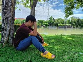 jeune homme asiatique aux cheveux noirs assis sous un arbre. stress et anxiété. à quoi tu penses dans la ville du parc d'été photo