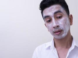 beau jeune homme asiatique appliquant de la crème sur son visage avec un visage souriant, concept de soins de la peau photo