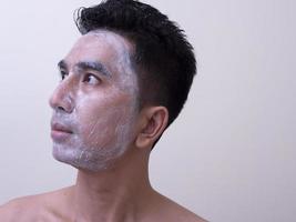 beau jeune homme asiatique appliquant de la crème sur son visage avec un visage souriant, concept de soins de la peau photo