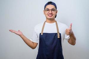 jeune et beau barista asiatique portant un tablier pointant quelque chose avec les mains, montrant le geste du pouce levé photo