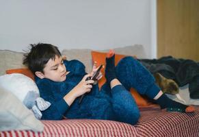 portrait cinématographique enfant heureux regardant un dessin animé sur une tablette, tir candide garçon mignon jouant à un jeu en ligne avec des amis sur internet, enfant confortable portant un pyjama moelleux assis sur un canapé se relaxant à la maison photo