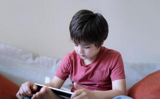 écolier assis sur un canapé en train de regarder un dessin animé sur une tablette, un jeune garçon mignon jouant à un jeu en ligne sur un pavé numérique, un portrait d'enfant cinématographique à l'intérieur s'amusant et se relaxant seul dans la salle d'étude photo