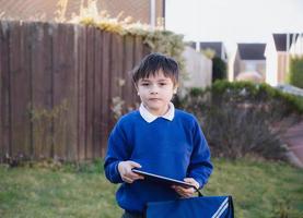 enfant heureux garçon tenant une tablette pc debout à l'extérieur en attendant le bus scolaire, portrait enfant jouant au jeu en ligne ou lisant l'histoire sur internet, garçon d'âge préscolaire apprenant avec la technologie moderne photo