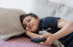 heureux jeune garçon tenant la télécommande et levant les yeux avec un visage souriant, mignon enfant allongé sur un canapé aime regarder une émission de télévision, enfant se relaxant à la maison le week-end photo