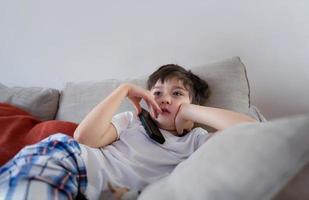 portrait jeune garçon au visage curieux assis sur un canapé en train de regarder un dessin animé, écolier assis seul sur un canapé tenant la télécommande en regardant la télévision, enfant se relaxant dans le salon après son retour de l'école. photo