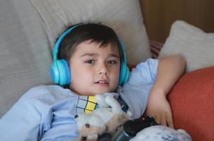 portrait d'enfant portant des écouteurs et jouant au jeu vidéo. enfant tenant une console de jeu en ligne avec des amis à la maison, jeune garçon assis sur un canapé s'amusant et se relaxant seul le week-end photo