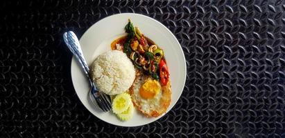 vue de dessus du basilic sauté avec calamars, riz, concombre tranché et œuf frit à la thaïlandaise sur plaque blanche avec espace de copie. mise à plat de plats célèbres et épicés sur fond noir en acier inoxydable. photo