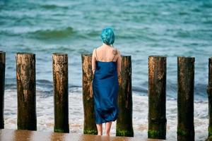 jeune femme aux cheveux bleus en longue robe bleu foncé debout sur une plage de sable regardant l'horizon de la mer photo