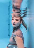 portrait d'art surréaliste de jeune femme en robe grise, écharpe perlée, petit sac, talons hauts violets sous l'eau dans la piscine photo