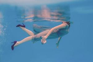 portrait d'art surréaliste de jeune femme en robe grise, écharpe perlée, petit sac, talons hauts violets sous l'eau dans la piscine photo