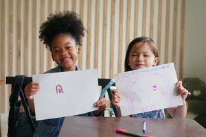 Dessin d'enfant d'âge préscolaire avec un crayon de couleur sur du papier blanc sur une table en classe avec des amis photo