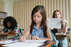 Dessin d'enfant d'âge préscolaire avec un crayon de couleur sur du papier blanc sur une table en classe avec des amis photo