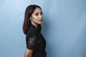 portrait latéral d'une femme asiatique séduisante, vêtue d'une chemise noire, souriant à la caméra, isolée par un fond bleu photo